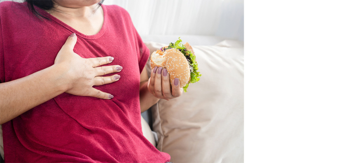 Übergewichtige Frau isst einen Burger und hat daraufhin mit Sodbrennen zu kämpfen.